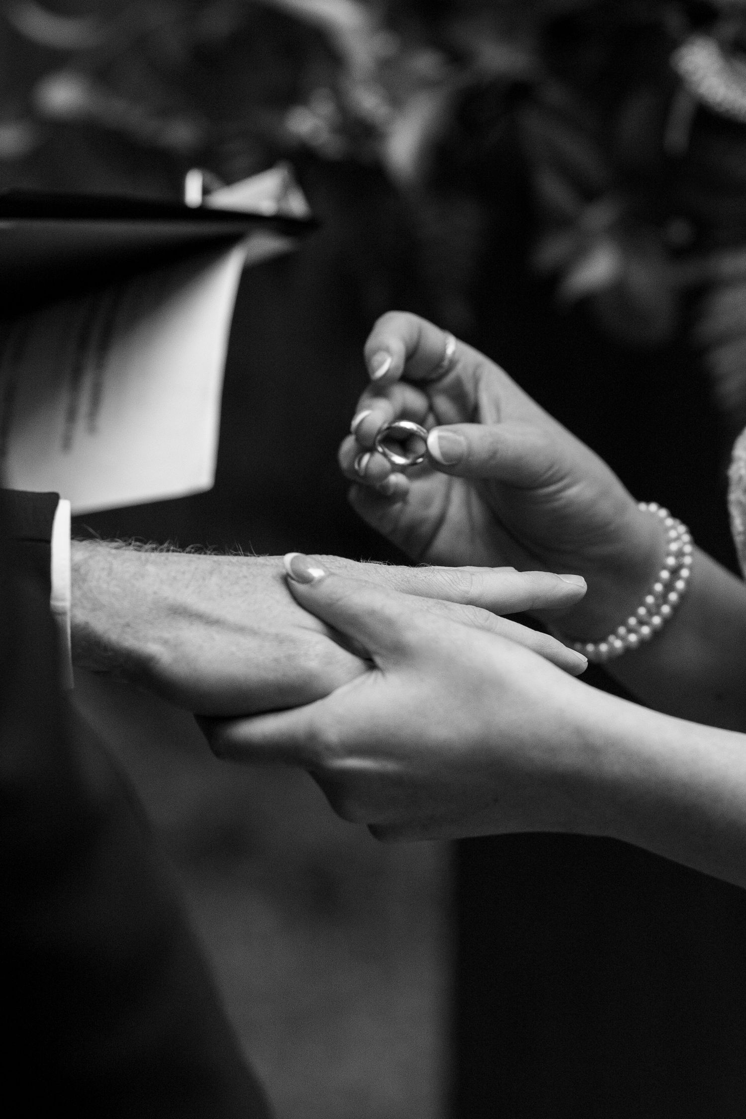 Black and white photo of a bride placing a wedding ring on the groom's finger during a ceremony.