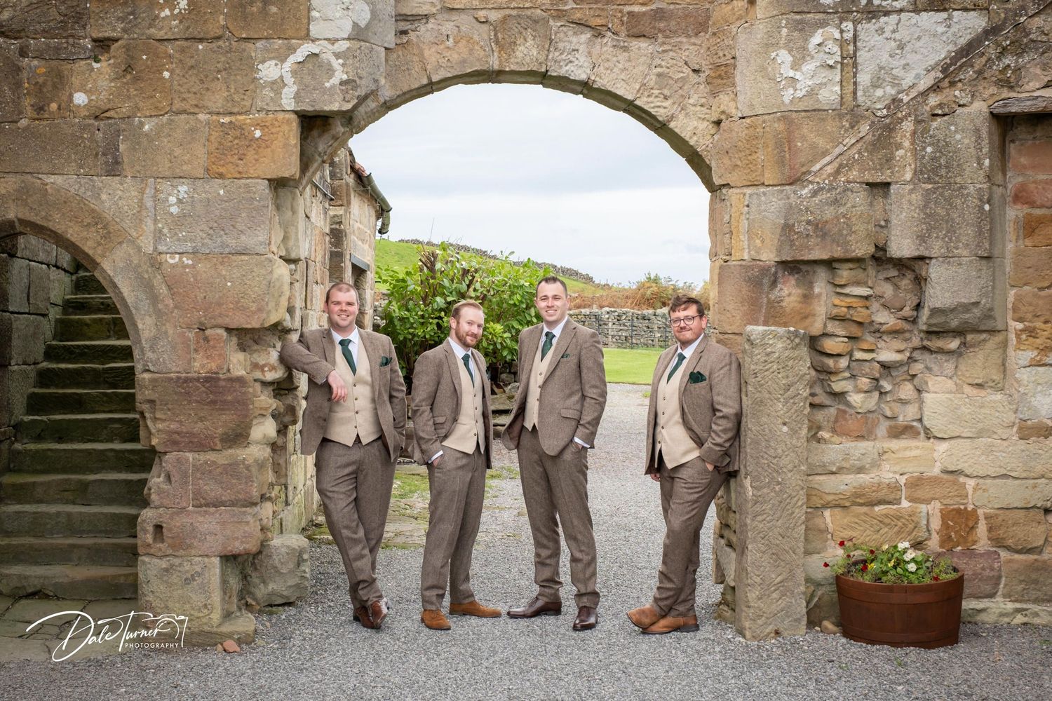 Groomsmen in coordinating brown tweed suits stand together under historic stone archways.