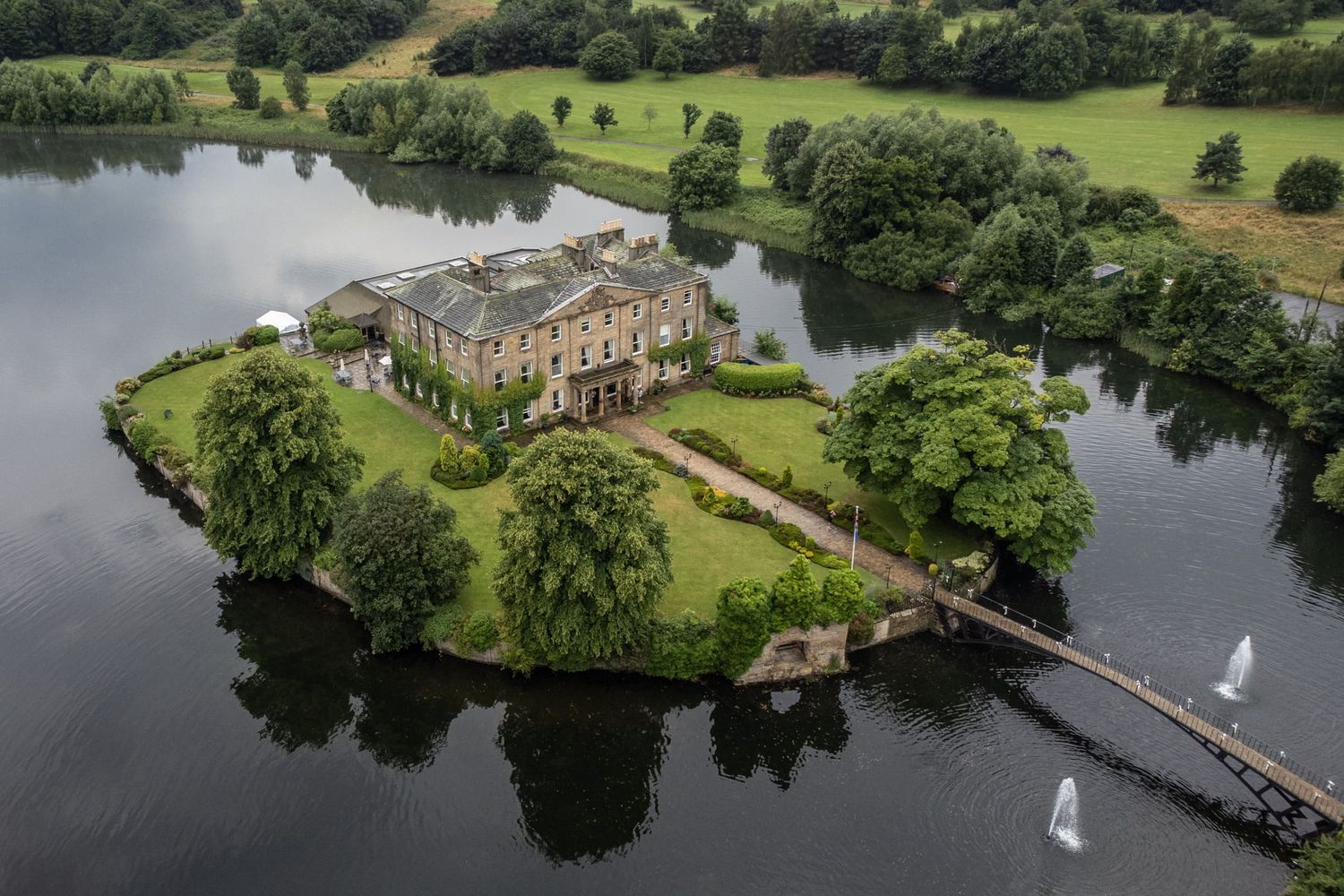 Aerial view of a grand mansion on a small island connected by a wooden bridge with sailboats on the lake.