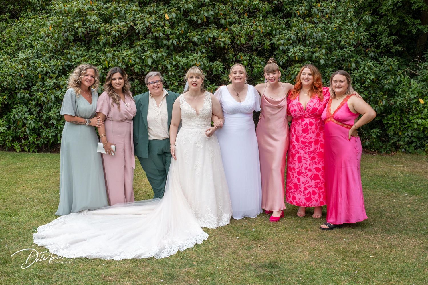 Wedding group photo session with mix of pink, white and green attire against natural backdrop.