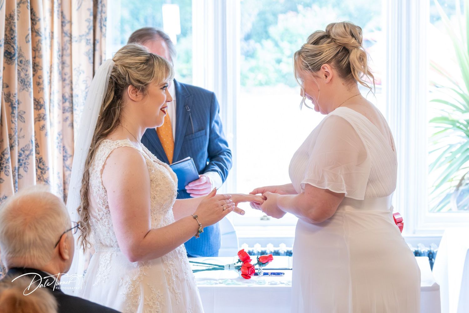 Wedding ceremony participants standing in a bright room with white curtains and natural lighting.