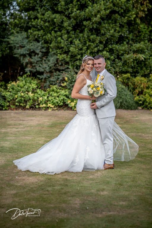 Newlyweds pose playfully on a grassy lawn with yellow bouquet and flowing white wedding dress.