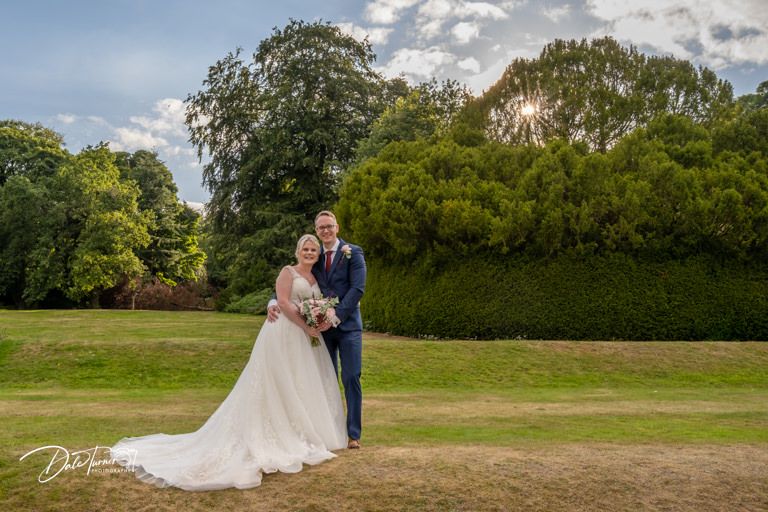 Wedding couple takes photos in a sunny field with cloudy sky backdrop.