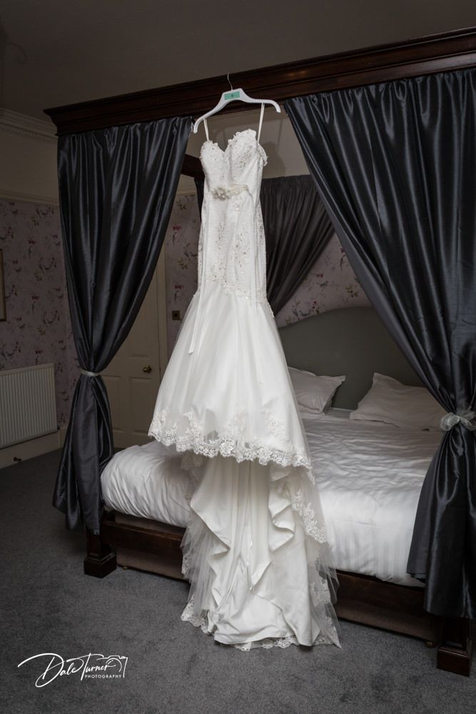 Elegant white lace wedding dress hanging in front of dark curtains in bedroom setting.