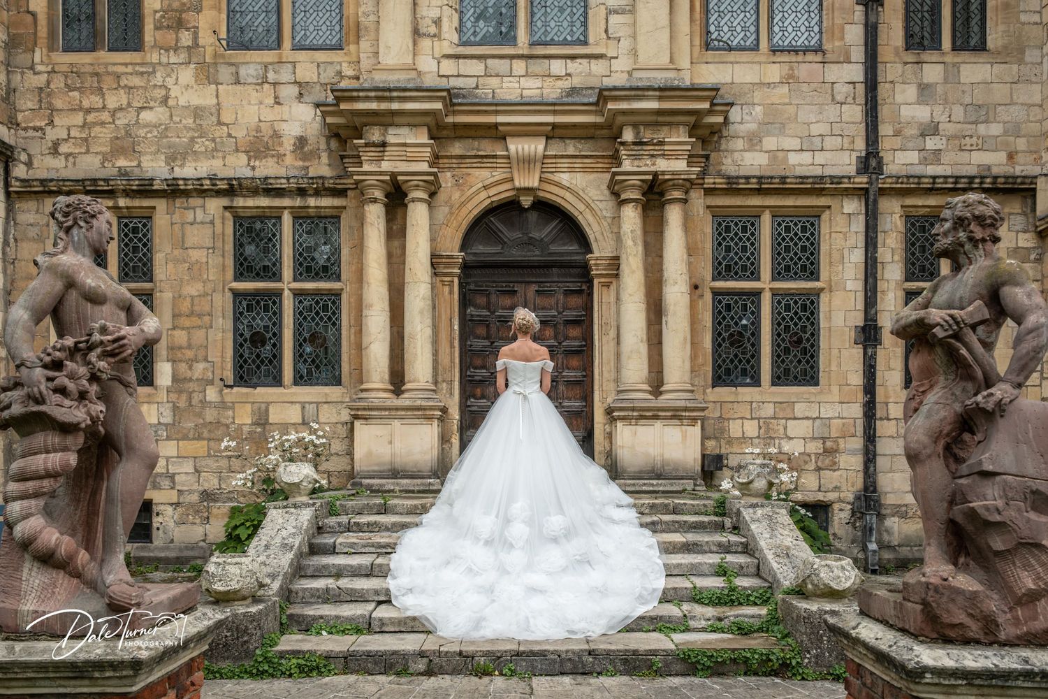 Bride in a stunning white ball gown stands on stone steps of a historic castle flanked by ornate statues.