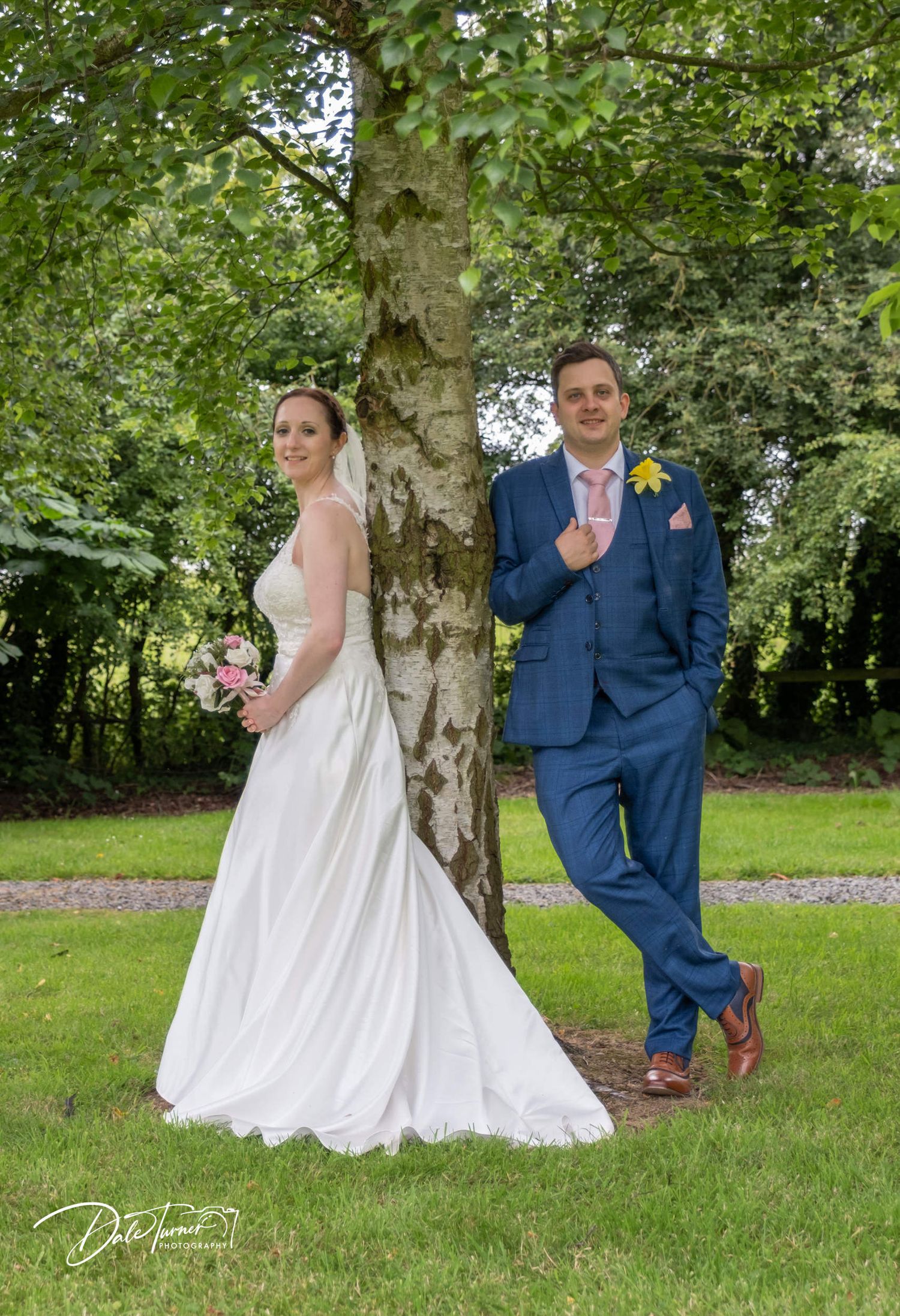 Couple poses playfully by tree trunk in garden wedding photoshoot.