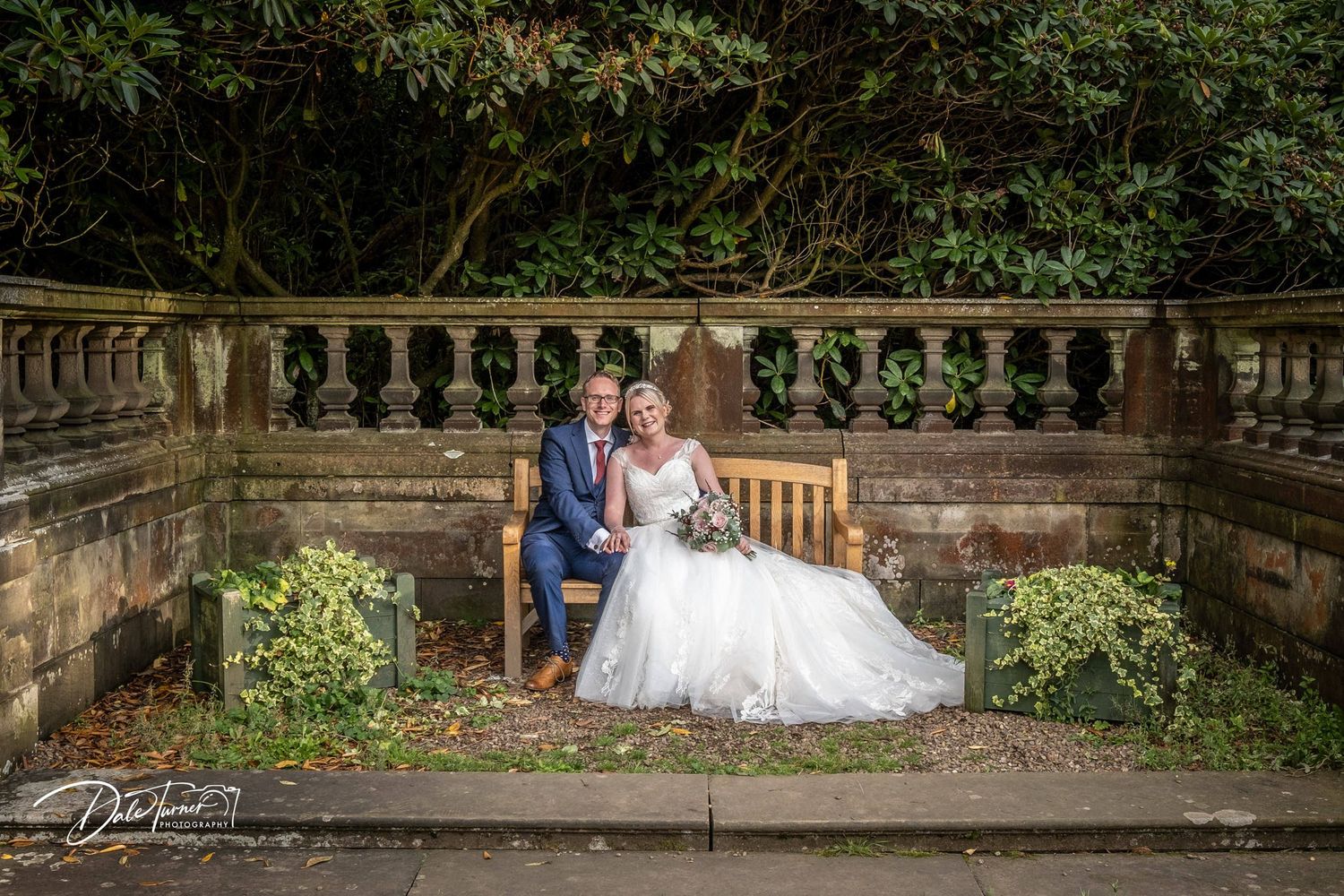 Wedding couple poses along a rustic stone wall with lush garden landscaping.