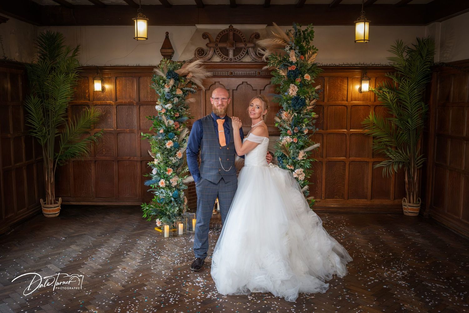 Bride and groom posing before a floral arch with tropical greenery in an elegant wood-paneled wedding venue.