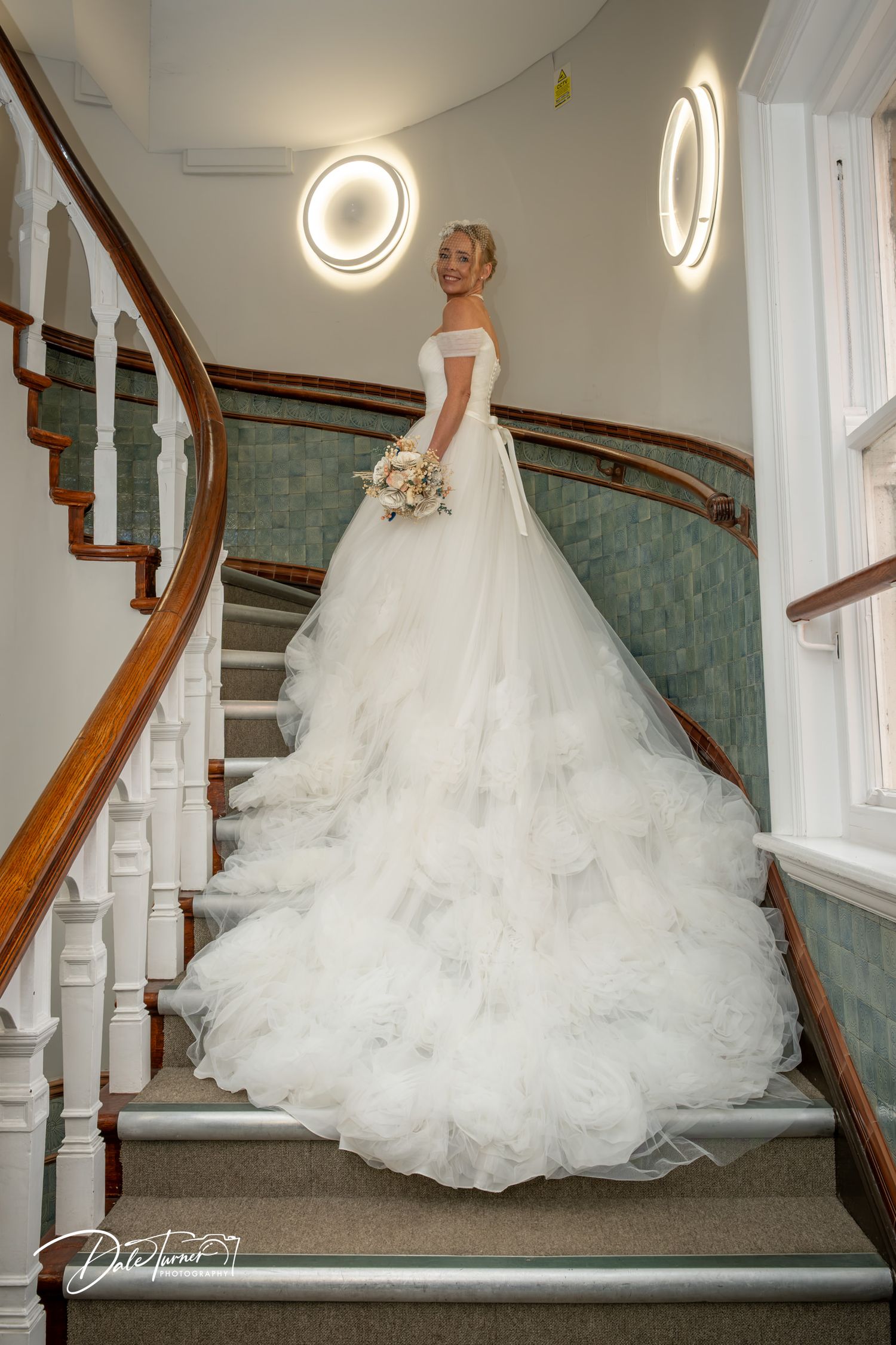 Bride in elegant white ballgown with long ruffled train poses on ornate wooden staircase holding bouquet.