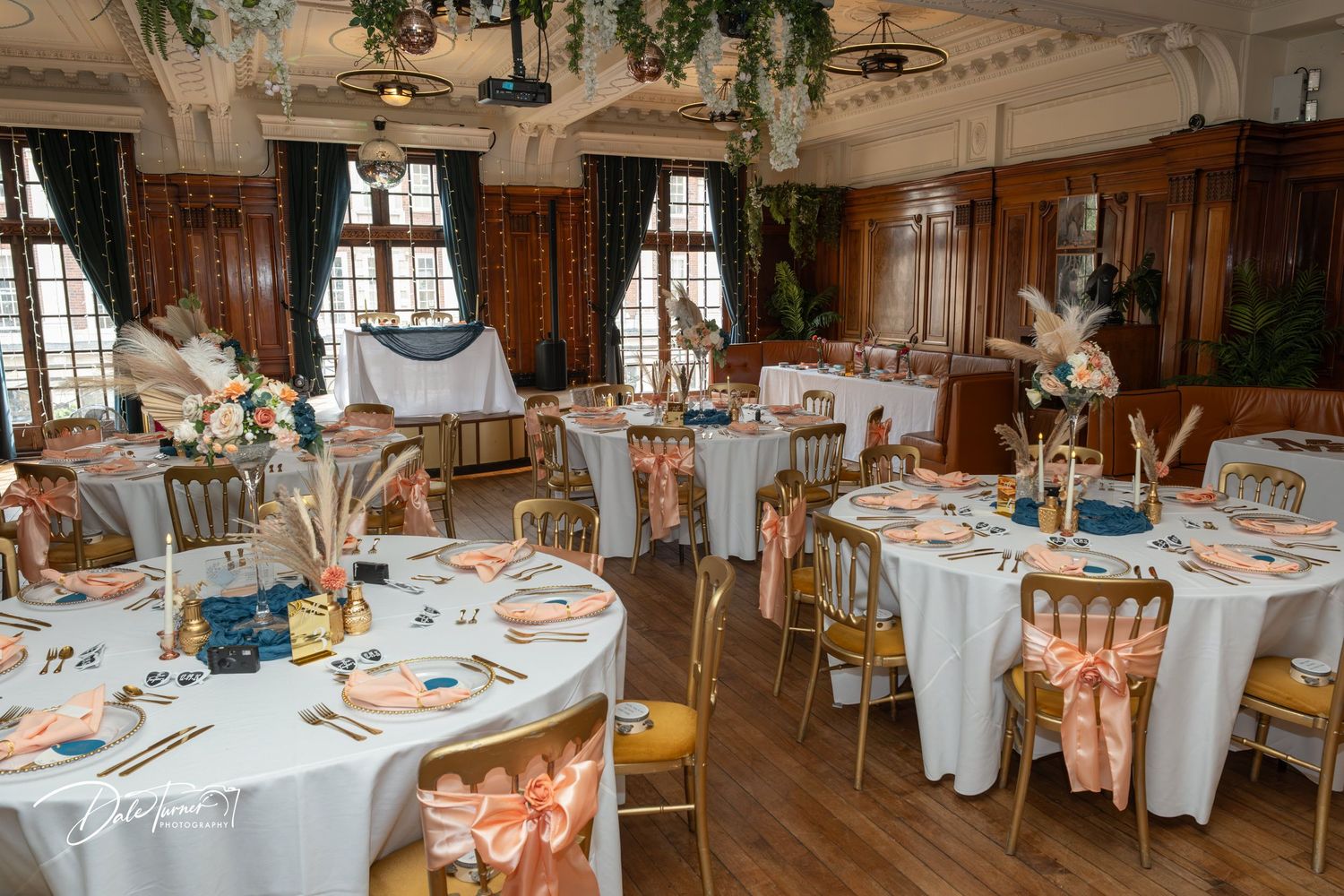 Elegant wedding reception hall with round tables, white linens, peach satin chair sashes, floral centerpieces, and warm wood paneling.