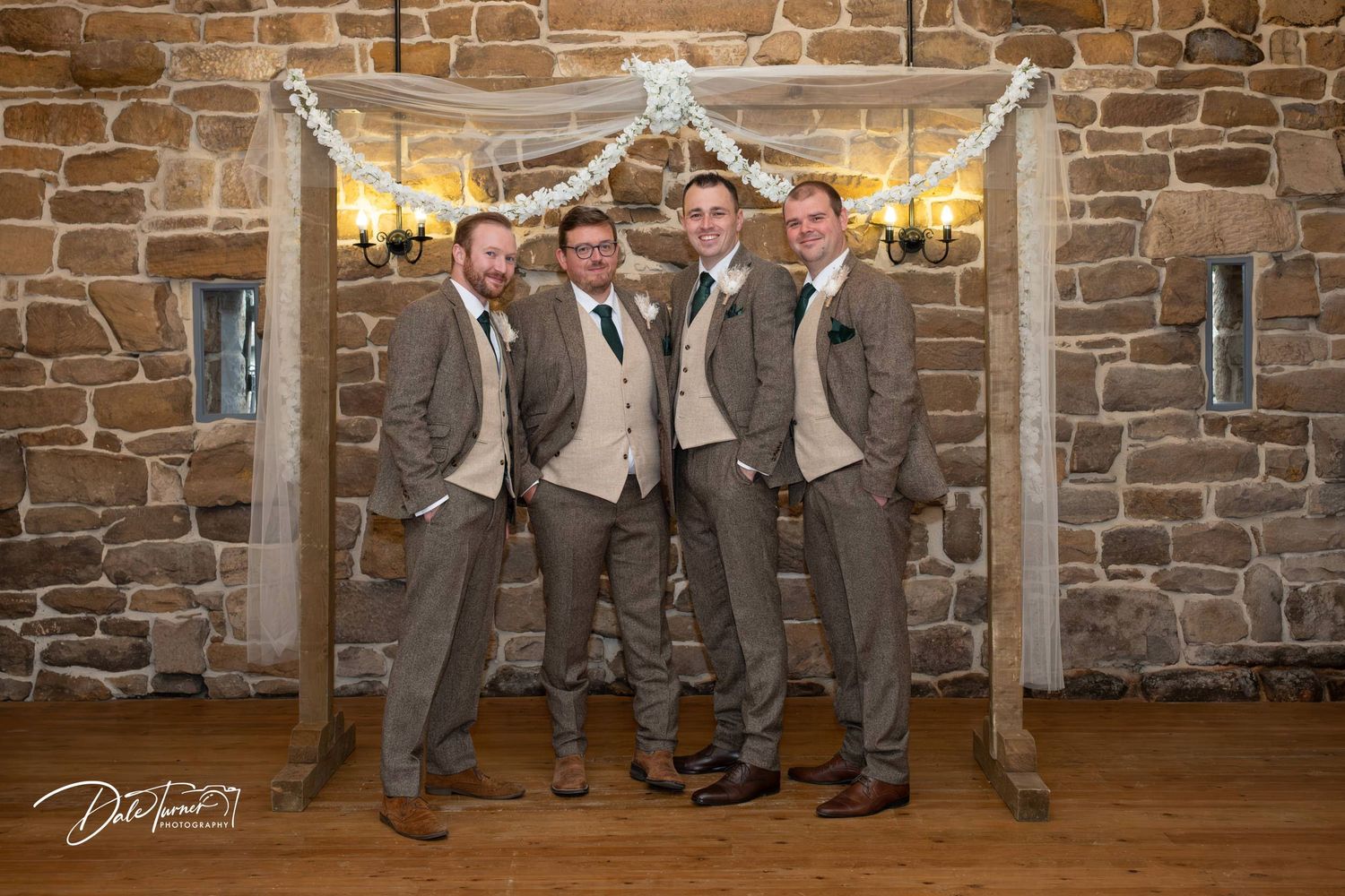 Groomsmen in gray suits pose playfully against rustic stone wall in wedding venue hallway.