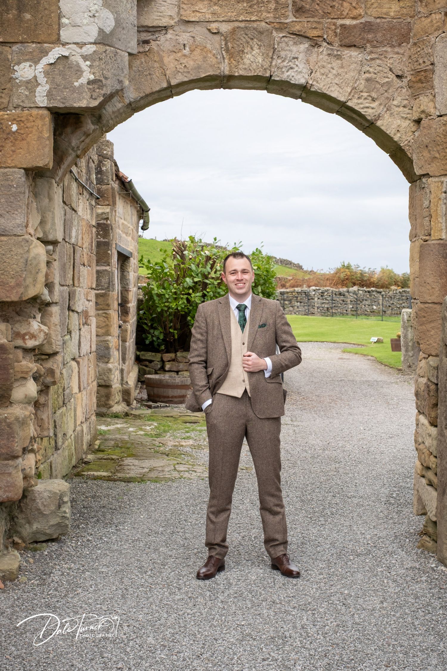 A wedding party member poses confidently under stone archways at a historic outdoor venue.