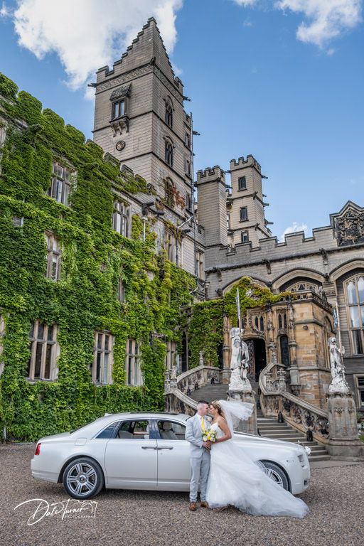 Classic white car parked in front of ivy-covered Gothic stone building.