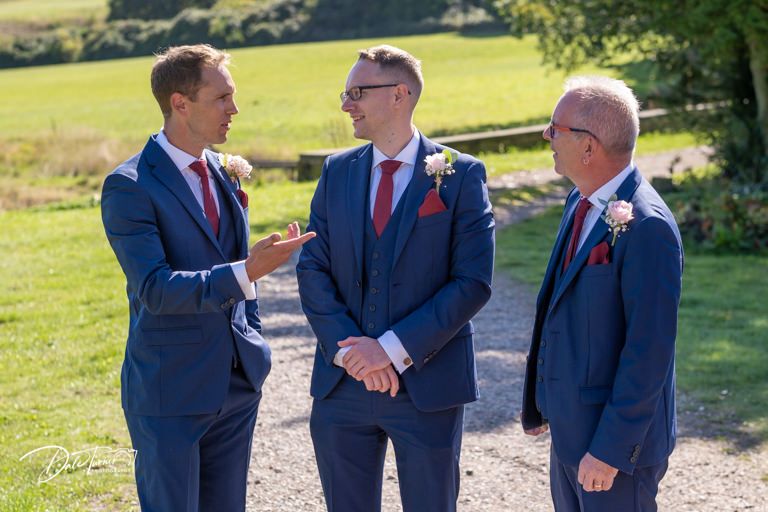 Three groomsmen in navy suits chatting outdoors on a sunny day.