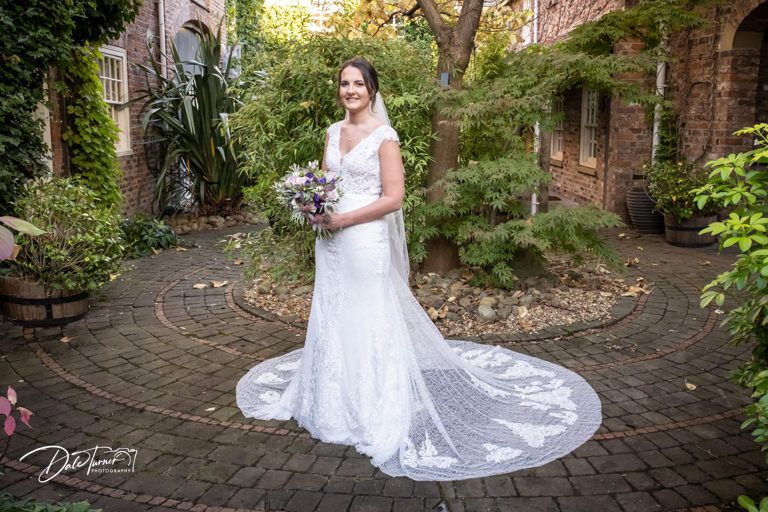 Bride in flowing lace gown poses in garden setting with historic brick wall backdrop.