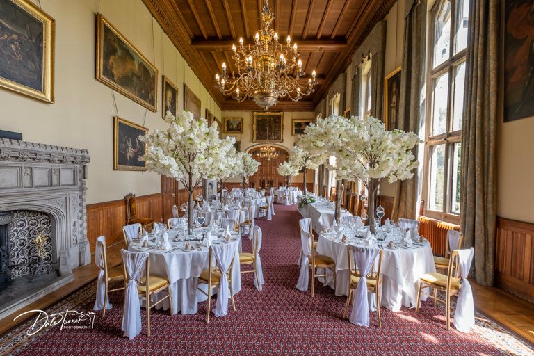 Grand banquet hall with crystal chandeliers and tall white floral arrangements on round tables.