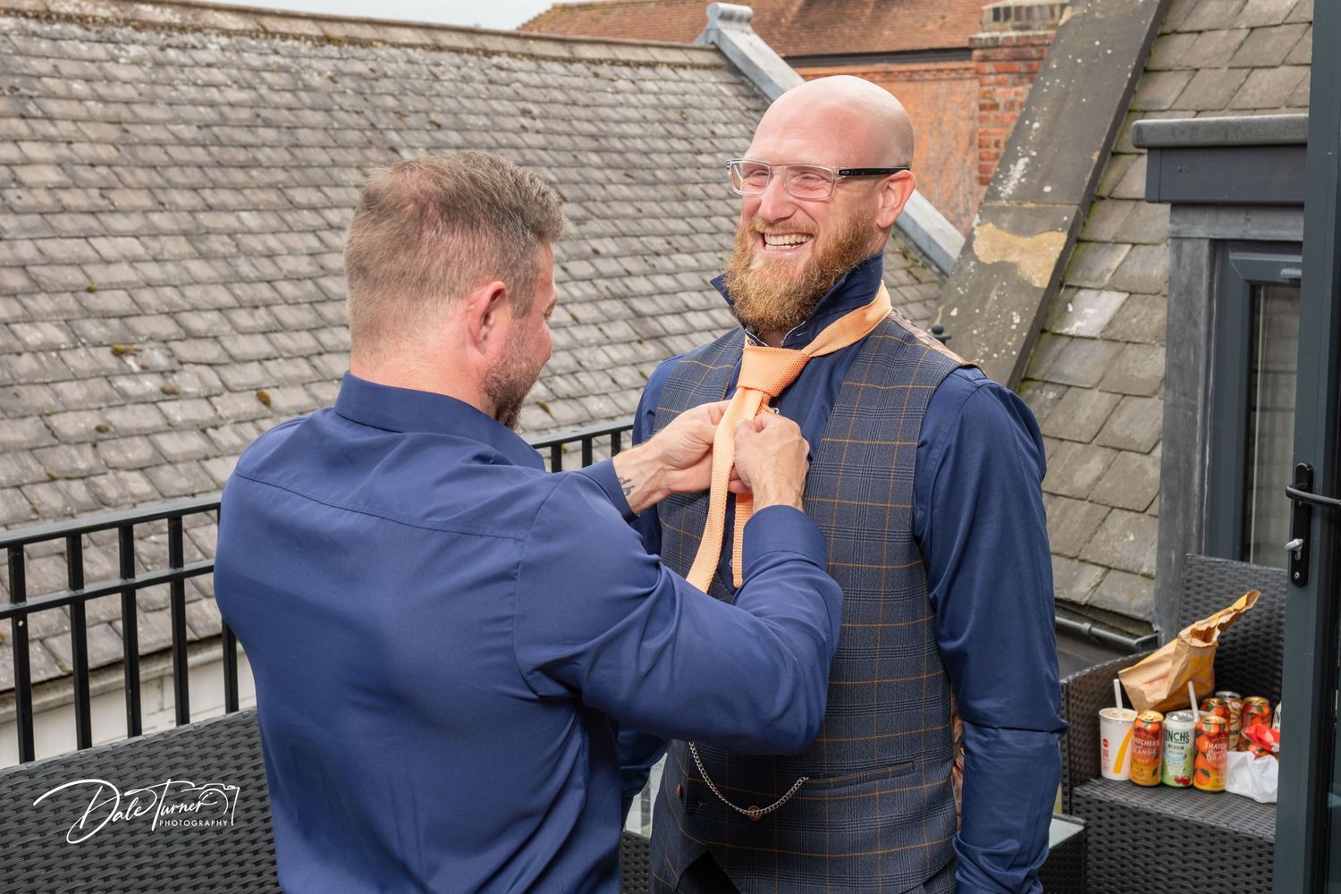A man helps a bearded groom adjust his orange tie outdoors on a rooftop terrace before a wedding ceremony.