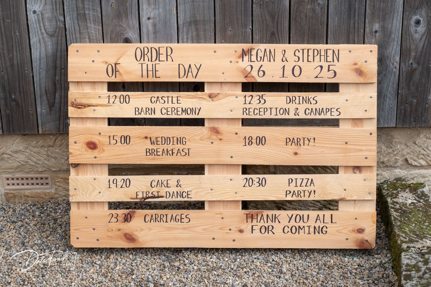 A rustic wooden pallet sign displays a wedding day timeline with various events and times written in chalk.