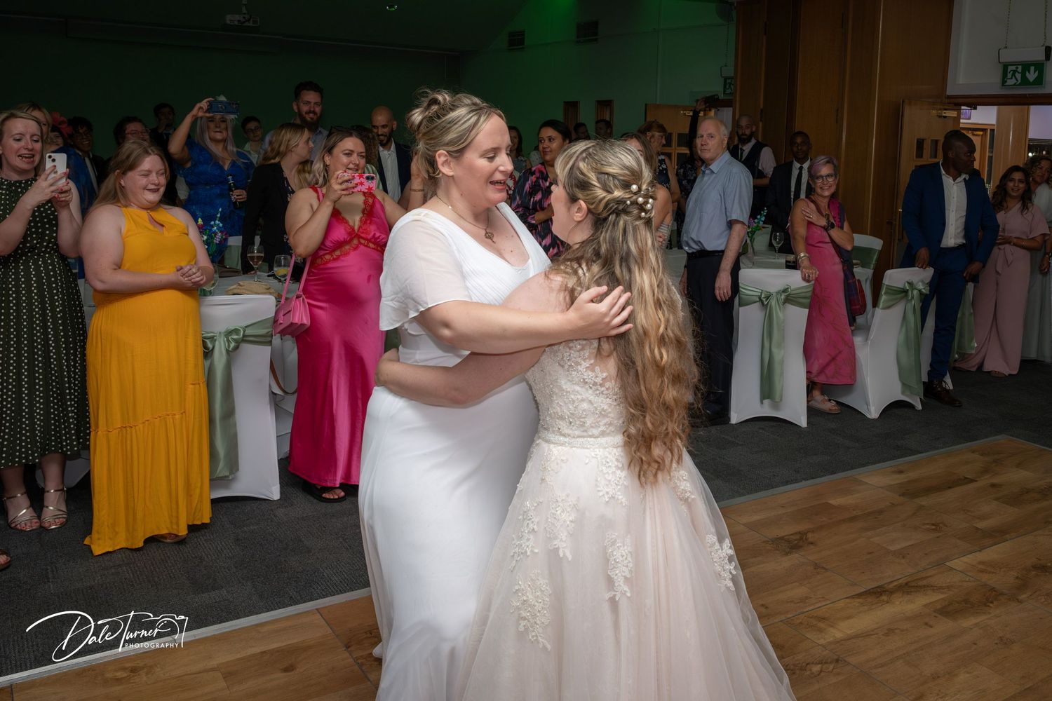 Two people in wedding attire share a joyful dance while guests in colorful dresses watch from the background.