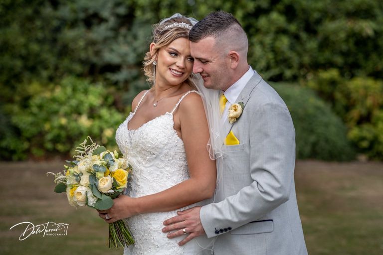 Wedding couple embracing outdoors in a garden setting with yellow floral bouquet details.