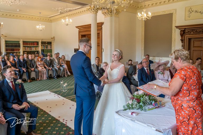 Formal wedding ceremony in progress inside historic venue with ornate chandeliers and carpeting.