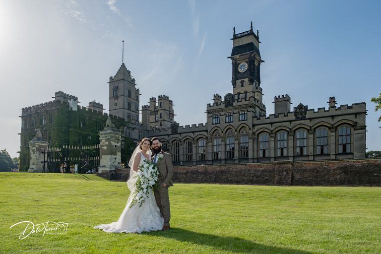 A wedding couple poses on the lawn in front of a grand historic castle with towers and ornate architecture.