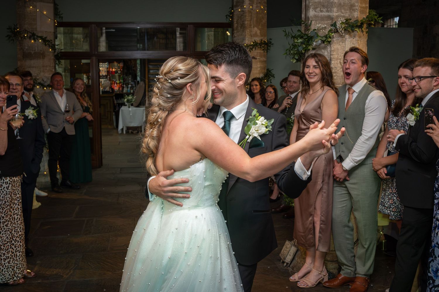 Newlyweds share their first dance surrounded by guests at a rustic indoor reception venue.