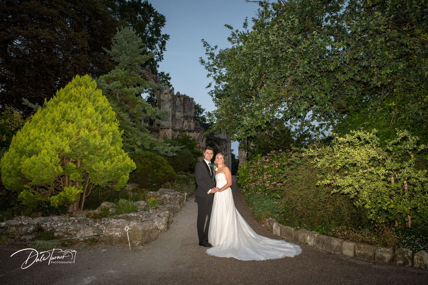 Romantic wedding portrait in a garden path with lush greenery and castle backdrop.
