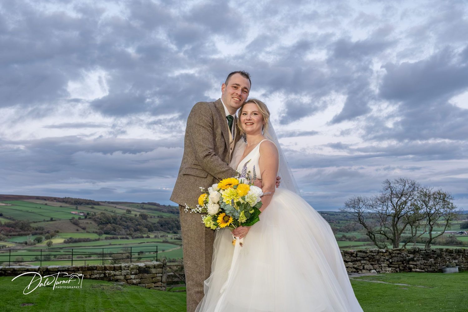 A couple shares a romantic moment in the Yorkshire countryside under dramatic cloudy skies with rolling hills in the background.