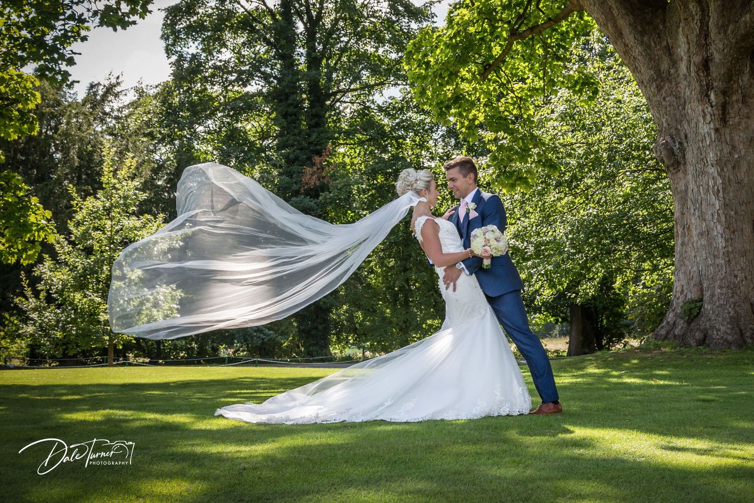 A wedding couple poses in a lush garden setting with a flowing veil catching the sunlight.