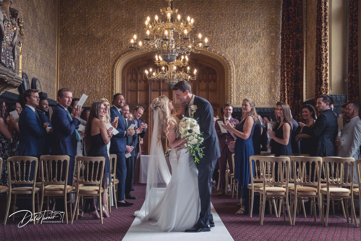 Wedding ceremony in grand ballroom with chandelier and gold chairs.