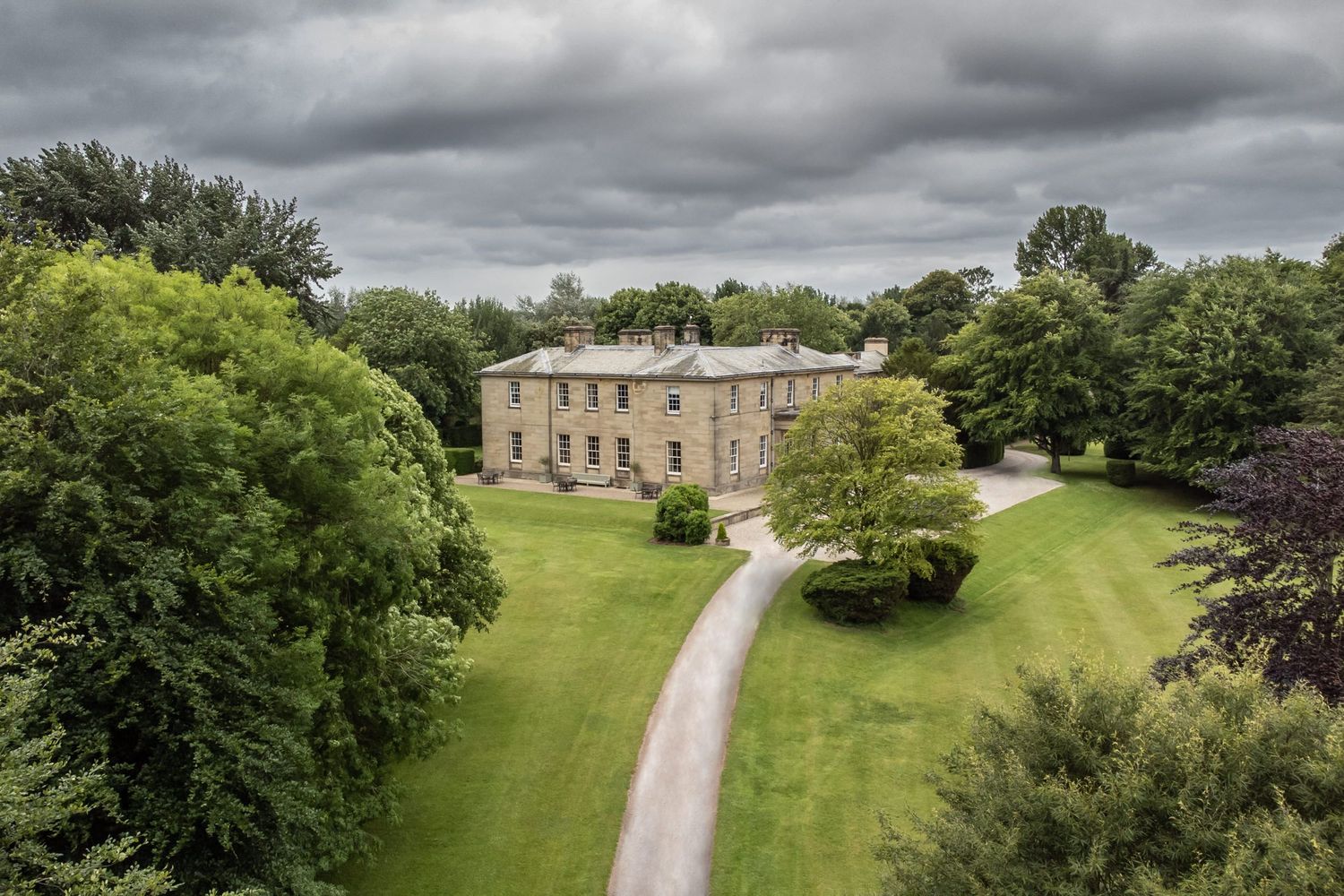 Aerial view of a stately home with winding driveway surrounded by manicured lawns and woodland.
