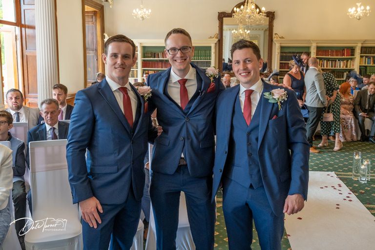 Groomsmen in navy suits with red ties pose together in an elegant room with chandeliers.
