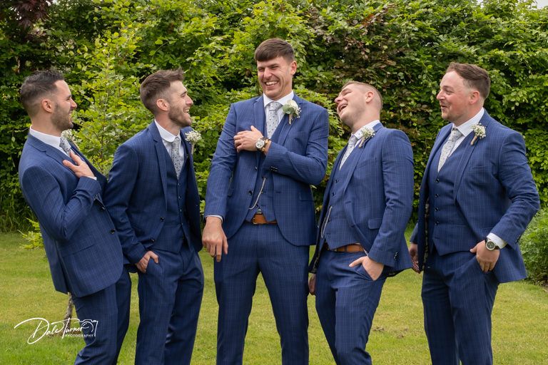 Groomsmen in navy blue suits share laughs while posing outdoors.