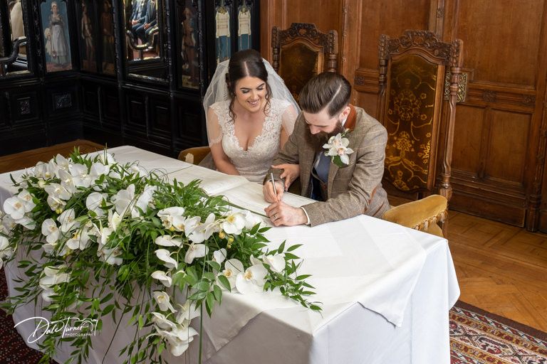 Wedding couple signing documents at white-clothed table with floral arrangements.
