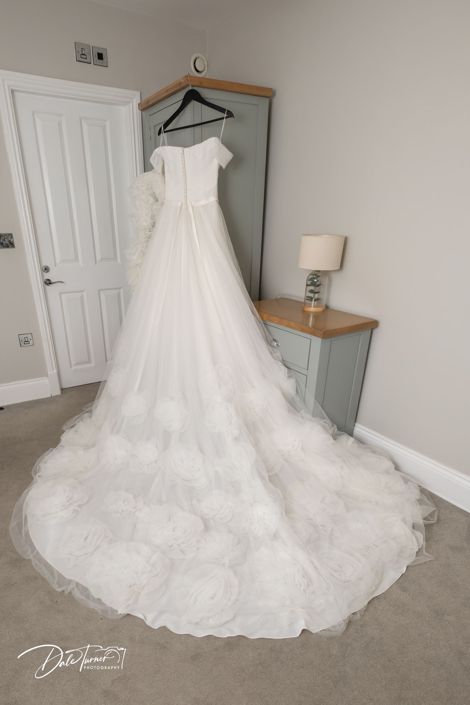 Elegant white wedding dress with ruffled train displayed on hanger in bridal suite room.