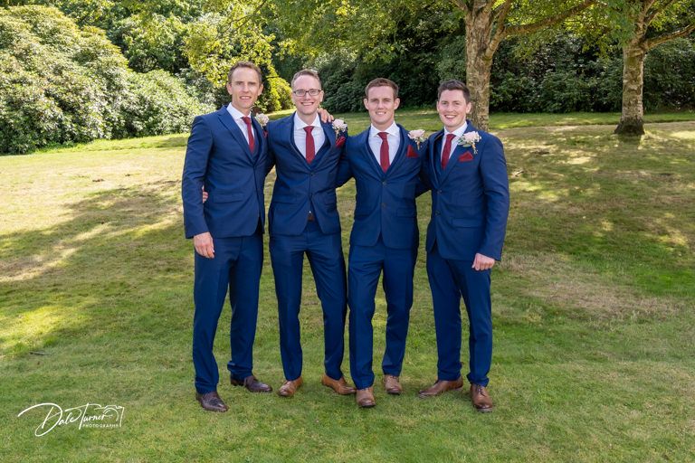 Groomsmen in navy blue suits pose together on a grassy lawn during a wedding celebration.