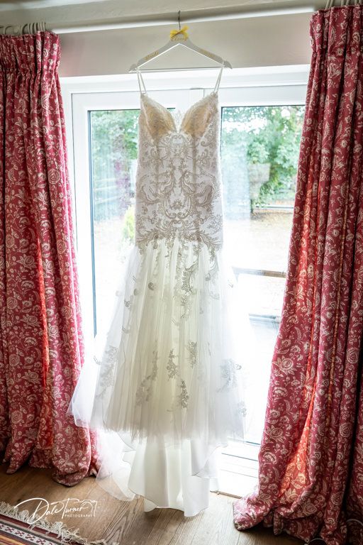 Wedding dress hanging by window with red floral curtains in morning light.