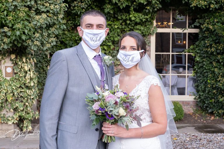 Wedding couple wearing protective masks pose together in garden setting with floral bouquet.