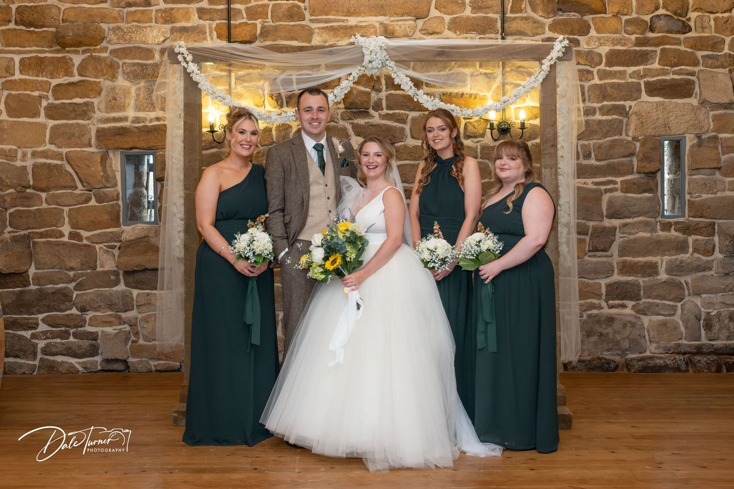 Wedding party poses in front of rustic stone wall with bridesmaids in dark green dresses and white bouquets.