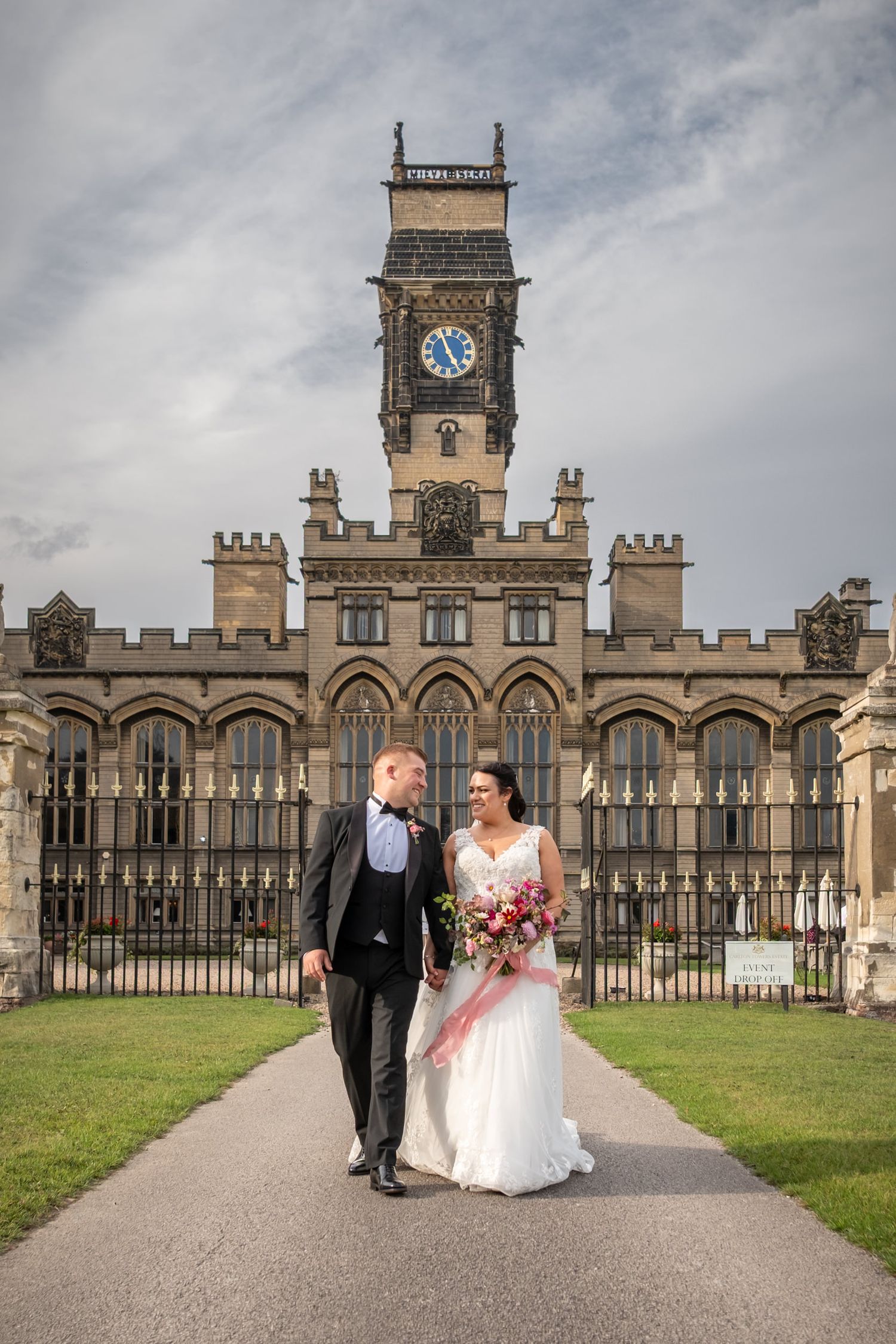 A wedding couple walks along a path in front of a grand historic building with a clock tower and ornate architecture.
