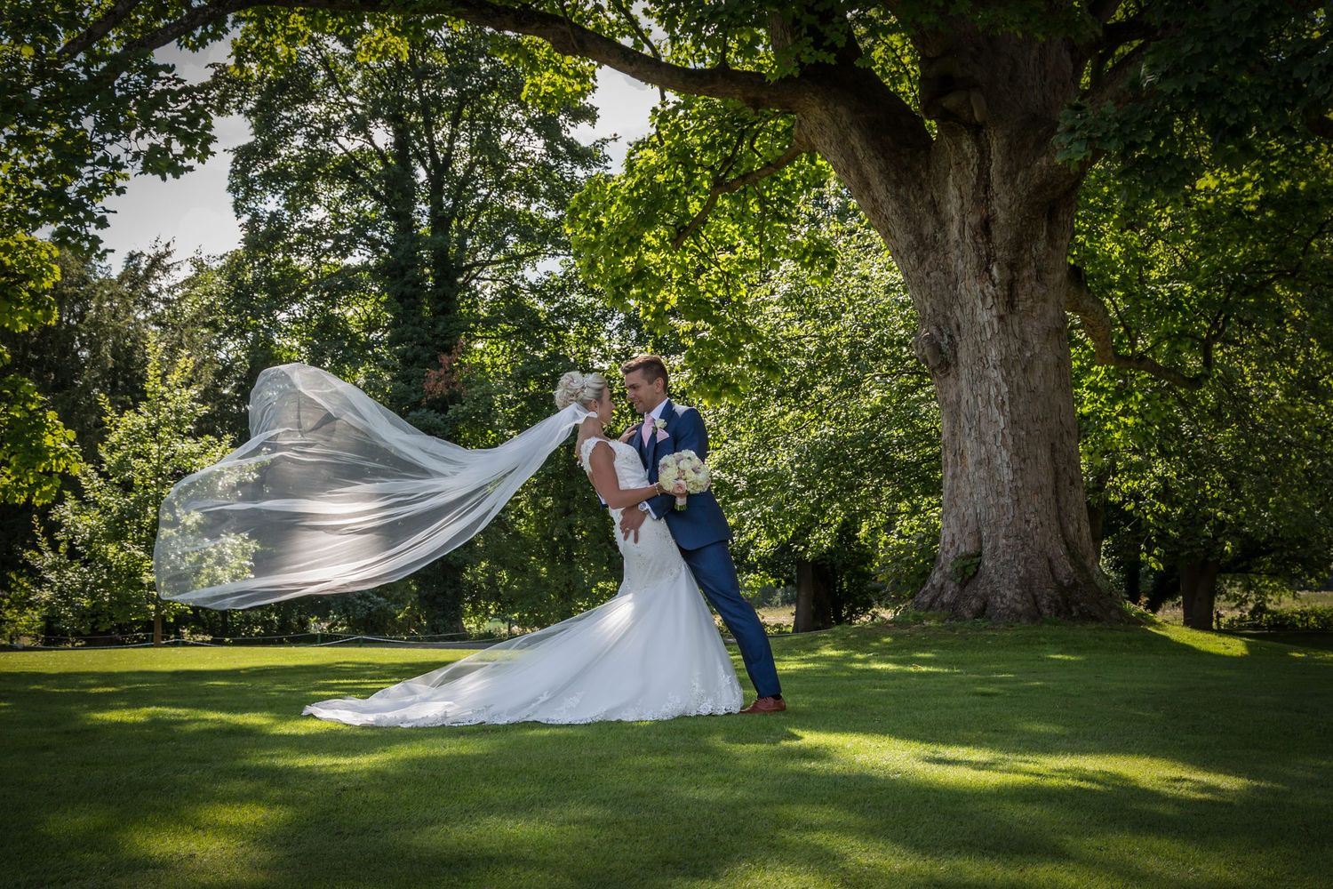 Wedding couple in garden setting with flowing veil caught in breeze under large tree.