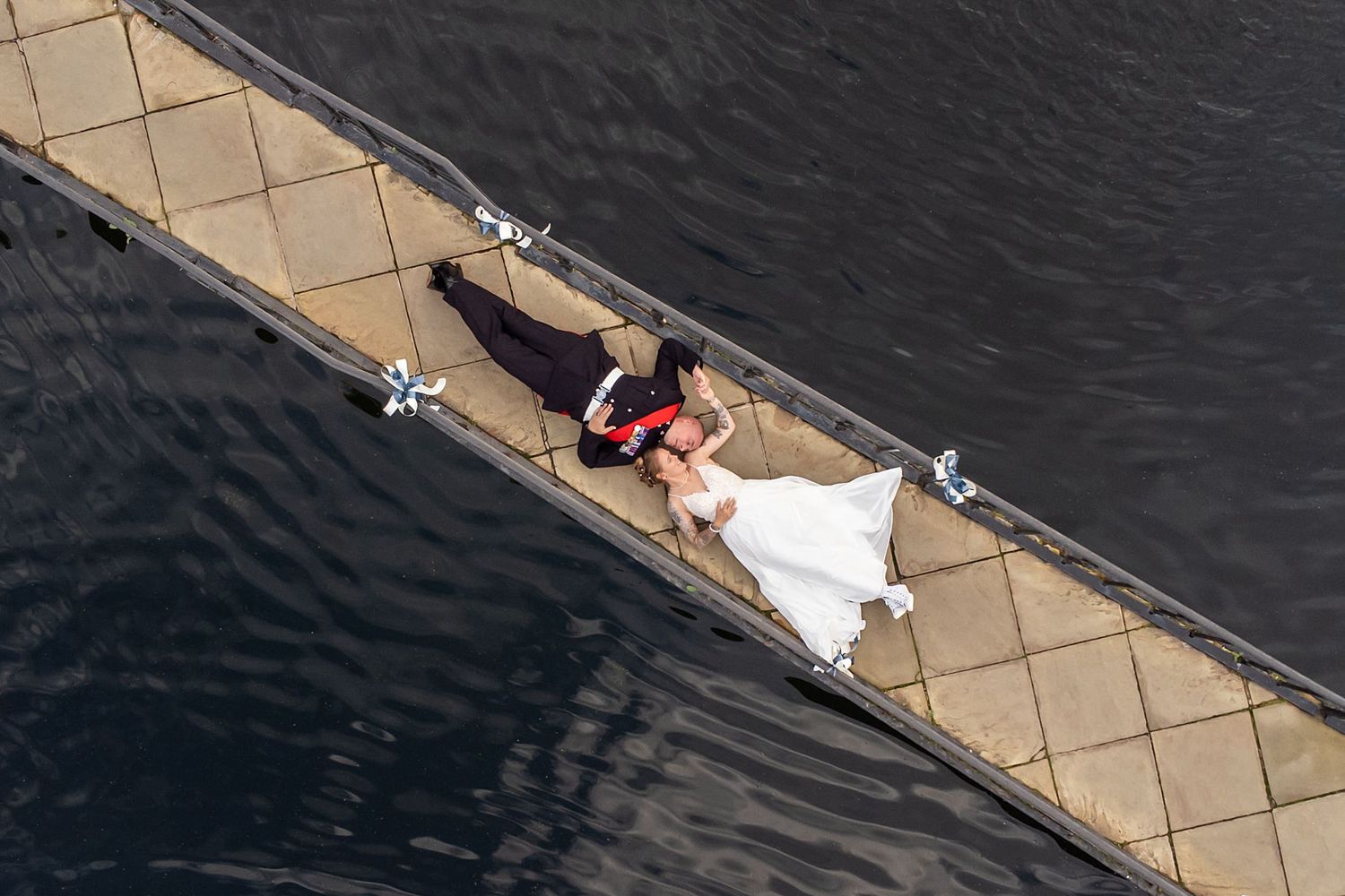 Aerial view of a couple in wedding attire lying on a stone pier by dark water.