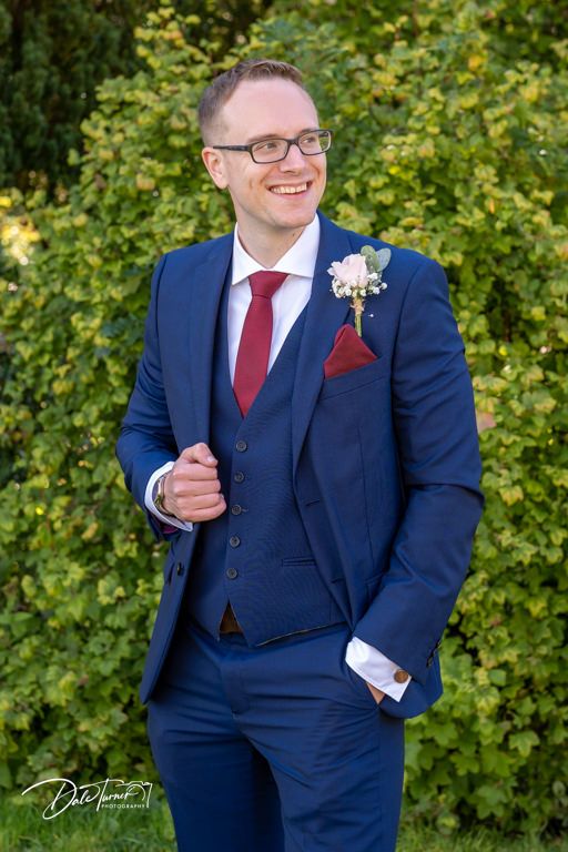 Groom in navy three-piece suit with red tie poses against green foliage.