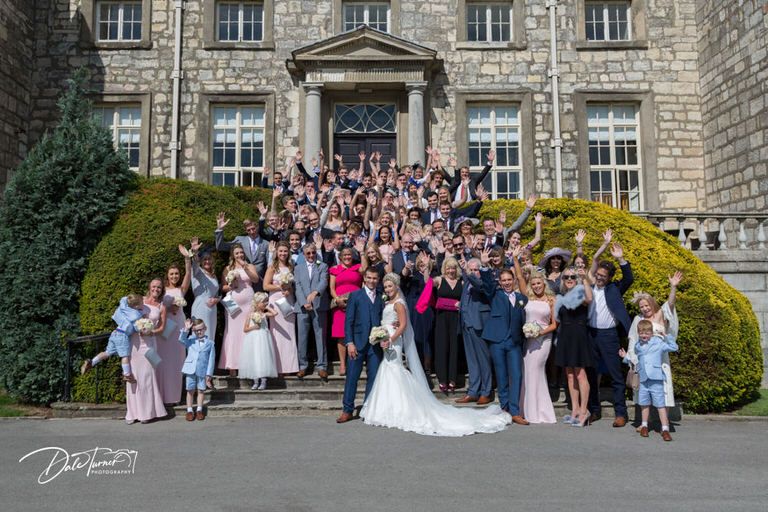 Large group wedding photo in front of historic stone building.