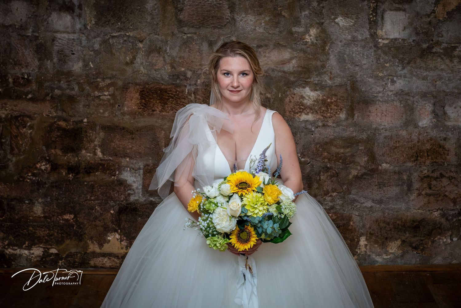 A rustic wedding scene with a white gown and sunflower bouquet against exposed brick walls.