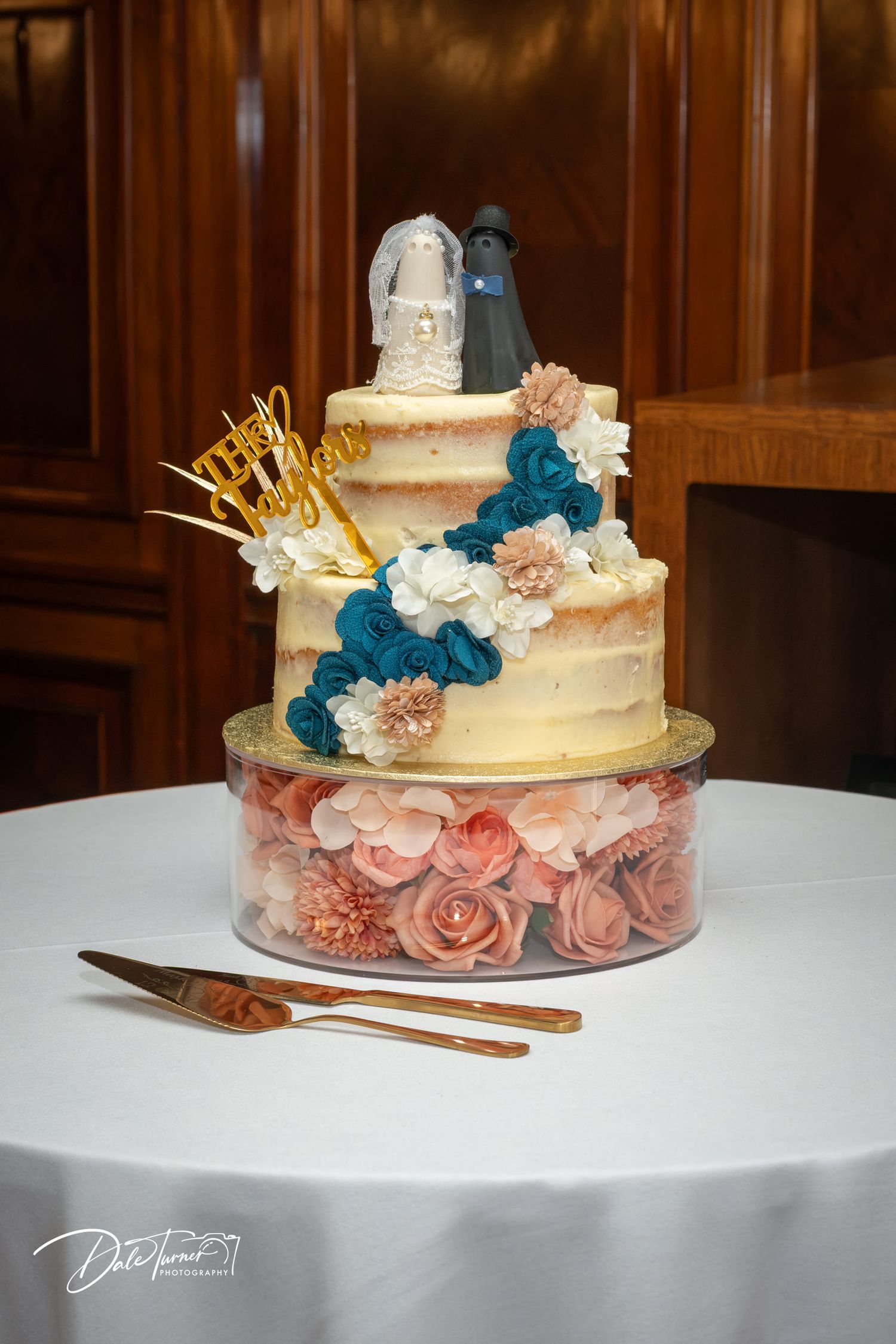 Three-tier semi-naked wedding cake with teal and pink flowers, bride and groom topper, and gold cake server set on white table.