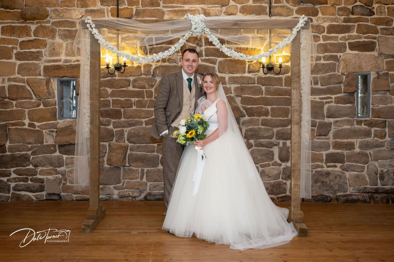 A wedding couple poses under a rustic burlap and light decorated arch against a stone wall backdrop.