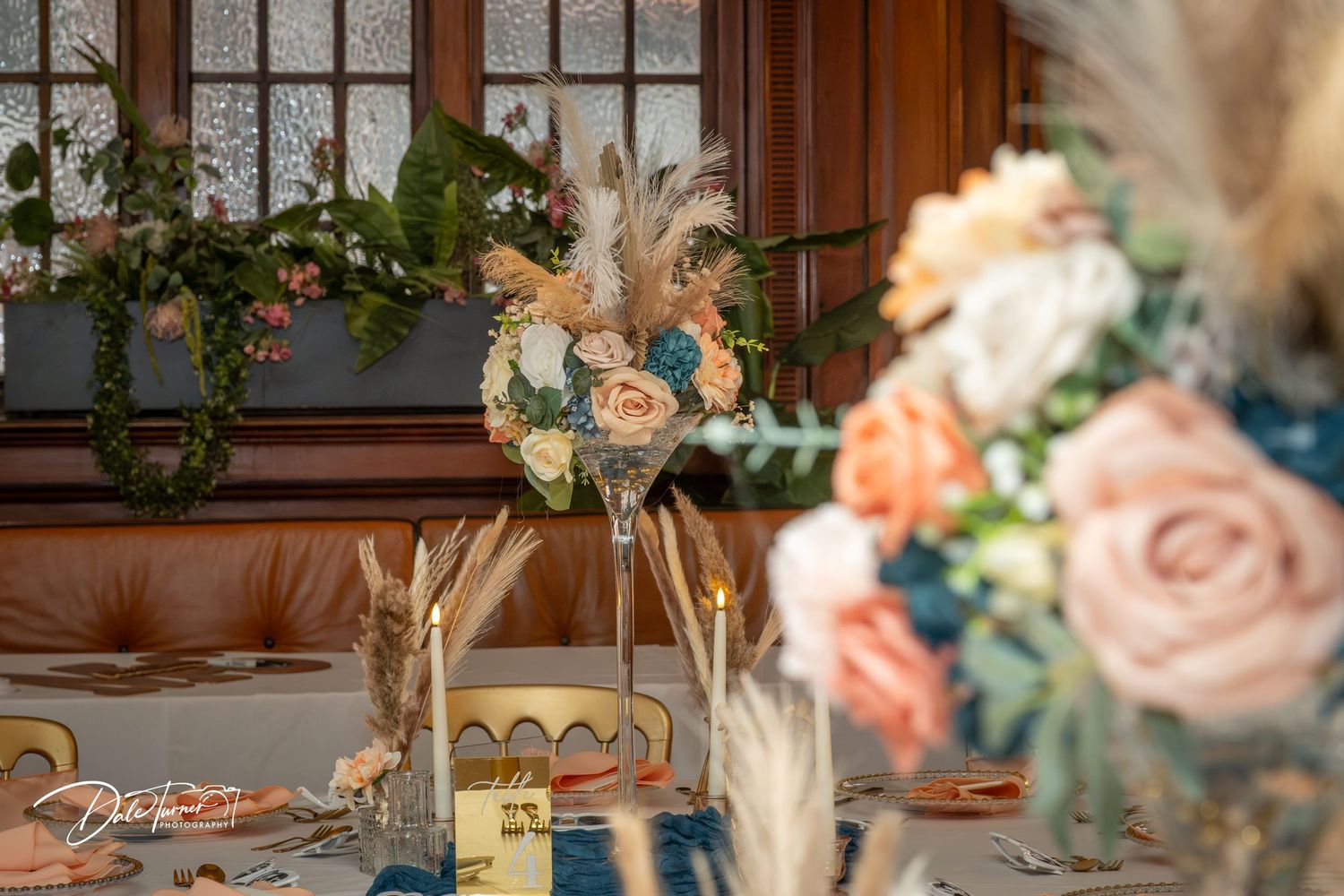 Elegant boho wedding table decor with feathered floral centerpiece, pampas grass, and peach roses in a warmly lit venue.