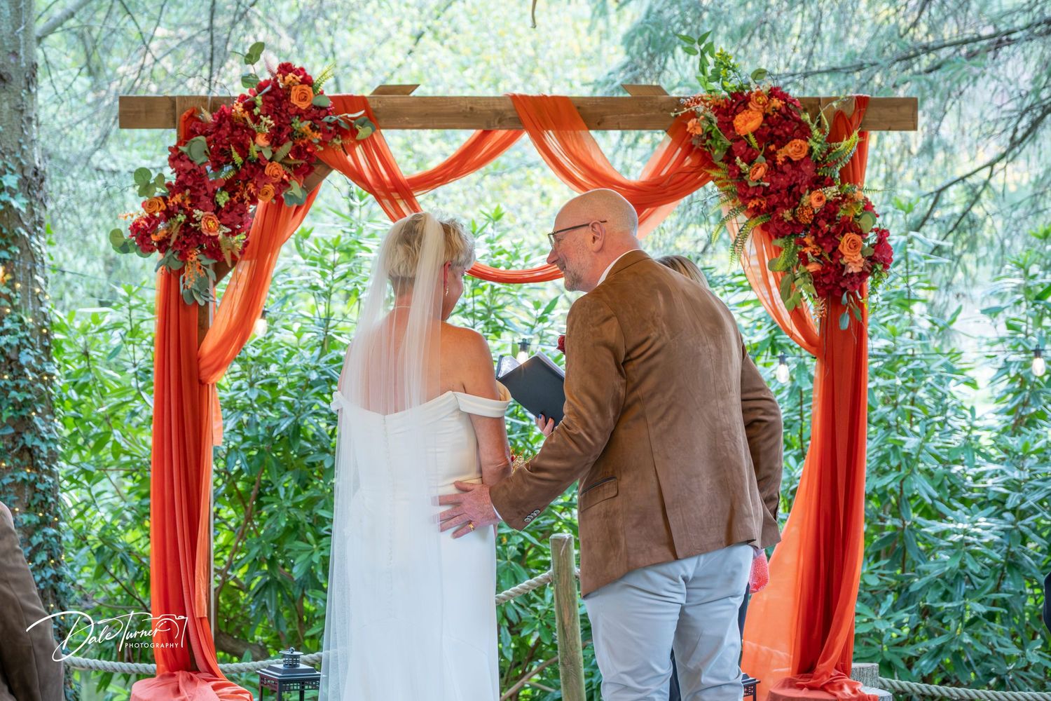 Wedding ceremony setup with red floral arch and outdoor garden backdrop.